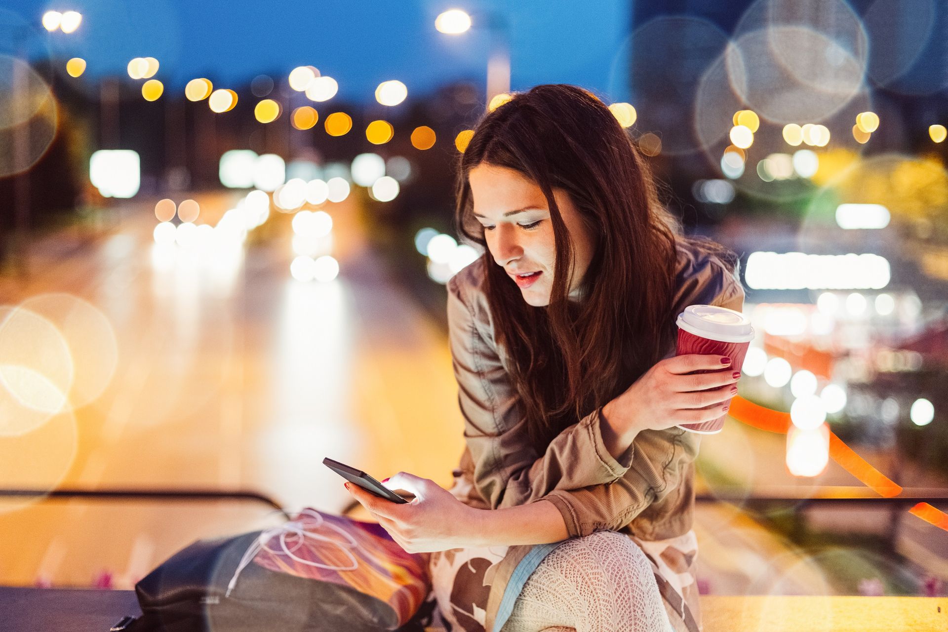 Frau sitzt auf einer Bank, hält ein Handy und eine Tasse Kaffee, lächelt in einer städtischen Umgebung.